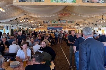 Blick in ein gut besuchtes Festzelt beim Eisenbahnertag; zahlreiche Besucherinnen und Besucher sitzen an Tischen oder stehen miteinander im Gespräch. Die Aufnahme zeigt das Innere eines großen Festzelts mit langen Tischreihen und vielen Gästen. Im Vordergrund sitzen mehrere Personen an Tischen mit Getränken und Geschirr. In der Mitte führt ein breiter Gang durch das Zelt, auf dem weitere Besucherinnen und Besucher stehen oder umhergehen. Im Hintergrund ist eine Bühne oder Veranstaltungsfläche mit einem Banner zu sehen, auf dem „Eisenbahnertag“ steht.