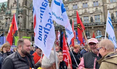 Gruppe von Menschen mit zahlreichen Fahnen vor der Fassade eines historischen Gebäudes. Mehrere weiß-blaue Fahnen mit teilweise sichtbarem Schriftzug „Verkehrs-“ sowie rote und schwarze Fahnen ragen in die Höhe. Im Vordergrund stehen mehrere Personen im Gespräch. Im Hintergrund sind weitere Transparente und Fahnen zwischen den Teilnehmenden zu sehen.