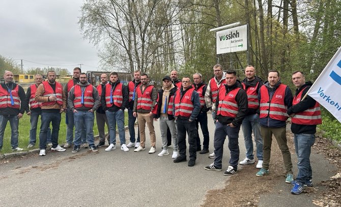 Eine Gruppe von Menschen in roten Warnwesten steht in einer Reihe an einer Straße neben Bäumen. Im Hintergrund ist ein Schild mit der Aufschrift „vossloh rail services“ zu sehen. Rechts am Bildrand ist eine Fahne mit EVG‑Logo erkennbar.
