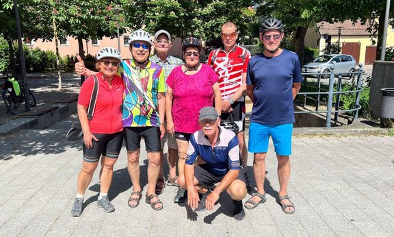 Acht Personen stehen und knien auf einem gepflasterten Platz im Freien, umgeben von Bäumen und Wohnhäusern. Mehrere Personen tragen Fahrradhelme und Sportkleidung, darunter bunte Trikots, T-Shirts, Shorts und Sandalen. Eine Person kniet vorne in der Mitte und trägt einen Fahrradhelm in der Hand. Hinter der Gruppe sind Fahrräder am Rand zu sehen sowie ein parkendes Auto und ein Geländer neben einer kleinen Vertiefung. Die Szene ist hell beleuchtet durch Sonnenlicht, mit deutlichen Schatten auf dem Boden.