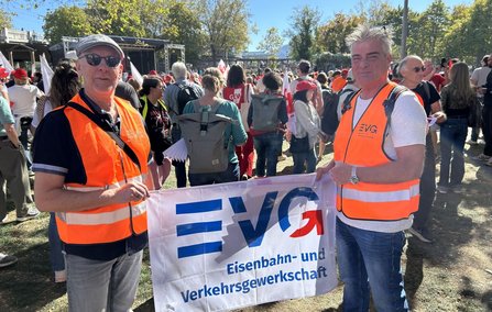 Zwei Personen mit orangefarbenen Warnwesten stehen im Vordergrund einer großen Versammlung im Freien und halten eine weiße Fahne mit der Aufschrift „EVG Eisenbahn- und Verkehrsgewerkschaft“. Im Hintergrund stehen zahlreiche Menschen, einige mit Fahnen, vor Bäumen und einer Bühne.