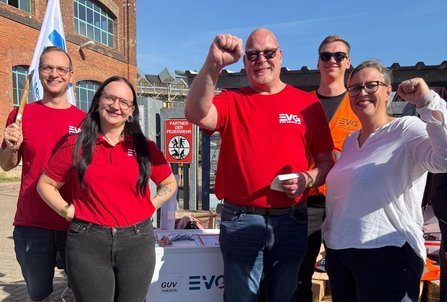 Fünf Personen stehen im Freien vor einem Tisch mit EVG-Logo. Zwei Personen tragen rote EVG-Shirts, eine weitere Person ein rotes EVG-T-Shirt, eine Person eine orangefarbene Warnweste mit EVG-Aufdruck, und eine Person eine weiße Bluse. Mehrere Personen heben eine geballte Faust. Im Hintergrund sind ein Backsteingebäude mit großen Fenstern, eine Fahne mit blauem Symbol sowie ein Schild mit der Aufschrift „PARTNER DER FEUERWEHR“ zu sehen. Der Himmel ist klar und blau.