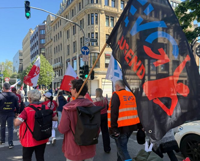 Rückansicht mehrerer Personen, die auf einer Straße gehen. Eine große schwarze Fahne mit dem Logo „EVG“ ist im Vordergrund. Eine Person trägt eine orange Warnweste mit „EVG“. Weitere rote Fahnen sind sichtbar. Im Hintergrund stehen mehrstöckige Gebäude, Bäume, Verkehrsschilder und eine Ampel.