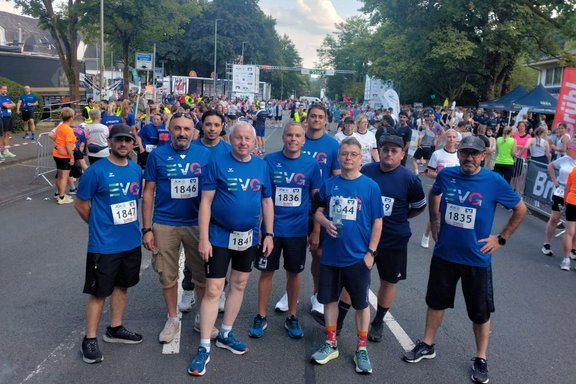 Neun Personen in blauen Laufshirts mit Startnummern stehen auf einer abgesperrten Straße vor einem Startbereich. Im Hintergrund befinden sich viele weitere Teilnehmende, Zuschauerbereiche mit Gittern, Bäume, Zelte und ein Startbanner über der Straße. Einige Personen tragen Sportkleidung und Laufschuhe, eine Person hält eine Wasserflasche.