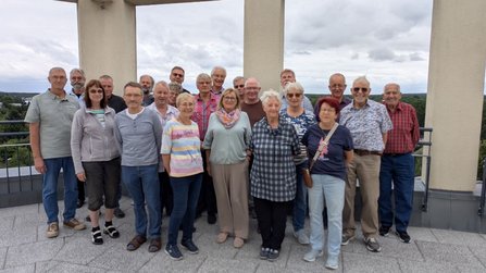 Gruppenfoto auf einer Dachterrasse oder Aussichtsplattform. Etwa 20 Personen stehen in mehreren Reihen vor einem Geländer. Im Hintergrund sind ein bewölkter Himmel und eine Landschaft mit Bäumen zu sehen.