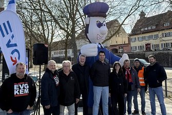 Eine Gruppe von neun Personen steht im Freien vor einem großen aufblasbaren Cartoon‑Figur in Uniform. Links im Bild befinden sich zwei Fahnen mit dem EVG‑Logo sowie technische Lautsprecher. Im Hintergrund sind kahle Bäume und mehrere Gebäude mit roten Dächern zu sehen.