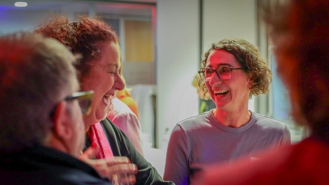 Mehrere Personen stehen in einem Innenraum zusammen. Im Vordergrund links ist der Hinterkopf einer Person zu sehen. Daneben steht eine Person mit lockigem Haar, Brille und einem hellen Oberteil. Links daneben steht eine weitere Person mit gebundenem Haar und einem dunklen Oberteil. Weitere Personen sind unscharf im Hintergrund erkennbar.