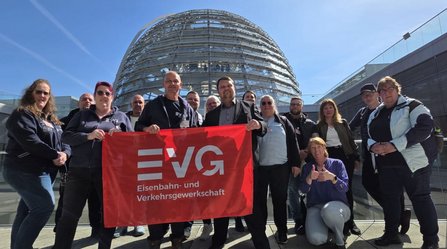 Gruppe von Personen steht vor einer großen gläsernen Kuppel eines Gebäudes. In der Mitte wird ein rotes Banner gehalten mit dem weißen Text: „EVG Eisenbahn- und Verkehrsgewerkschaft“. Die Personen stehen auf einer Terrasse mit Glasgeländer, blauer Himmel im Hintergrund.