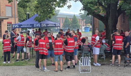 Mehrere Personen in roten Warnwesten mit der Aufschrift „STREIK!“ stehen und sitzen im Freien unter Bäumen. Im Hintergrund befindet sich ein blaues Zelt mit EVG-Logos und dem Schriftzug „Eisenbahn- und Verkehrsgewerkschaft“. Daneben sind Tische, Stühle und weitere Personen zu sehen. Backsteingebäude befinden sich im Hintergrund.