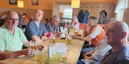 Mehrere Personen sitzen an einem rechteckigen Holztisch. Auf dem Tisch stehen Getränkegläser, Bierkrüge, Flaschen und kleine Vasen mit Blumen. Im Hintergrund sind weitere Sitzplätze, Wandhaken und Fenster zu sehen.