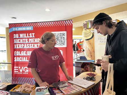 Informationsstand in einem Innenraum. Vor einem roten Aufsteller mit der Aufschrift „EVG JUGEND NRW“, einem QR-Code und dem Text „DU WILLST NICHTS MEHR VERPASSEN? FOLGE UNS AUF SOCIAL MEDIA!“ steht eine Person hinter einem Stehtisch. Auf dem Tisch liegen Broschüren, Flyer, Schlüsselanhänger, Stofftaschen und Informationsblätter. Eine weitere Person steht gegenüber und blickt auf ein Smartphone. Im Hintergrund sind weitere Stände und eine große dekorative Figur zu sehen.