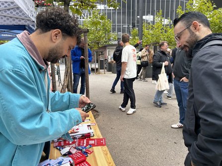 Außenbereich vor einem Gebäude mit Glasfassade und Bäumen. Im Vordergrund befindet sich ein Holztisch mit Flyern, Werbematerial und Süßigkeiten. Eine Person hält ein Smartphone über dem Tisch. Mehrere Personen stehen oder gehen auf dem Platz. Links ist ein blaues Zelt teilweise sichtbar.
