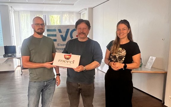 Drei Personen stehen in einem hellen Raum vor einem Banner mit dem teilweise sichtbaren Text „EVG Eisenbahn- und Verkehrsgewerkschaft“. Die Person in der Mitte hält gemeinsam mit der Person links eine weiße Schachtel mit dem Aufdruck „merci“. Im Hintergrund sind Fenster, Tische und ein Monitor zu sehen.