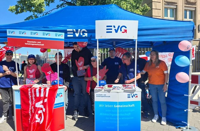 Blauer Pavillon im Freien mit zwei weißen Stehtischen. Auf dem Pavillon und den Tischen steht das Logo „EVG“. Auf einem Schild steht „Eisenbahn- und Verkehrsgewerkschaft“. Mehrere Personen stehen hinter und neben den Tischen und halten rote Pappschilder mit einer stilisierten Hand und Herz. Auf den Tischen liegen Flyer und kleine Gegenstände. Rechts sind bunte Luftballons befestigt. Im Hintergrund sind Bäume und ein Gebäude zu sehen.