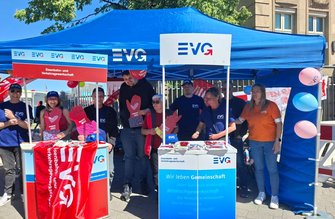 Blauer Pavillon im Freien mit zwei weißen Stehtischen. Auf dem Pavillon und den Tischen steht das Logo „EVG“. Auf einem Schild steht „Eisenbahn- und Verkehrsgewerkschaft“. Mehrere Personen stehen hinter und neben den Tischen und halten rote Pappschilder mit einer stilisierten Hand und Herz. Auf den Tischen liegen Flyer und kleine Gegenstände. Rechts sind bunte Luftballons befestigt. Im Hintergrund sind Bäume und ein Gebäude zu sehen.