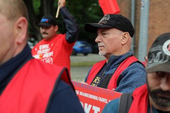 Nahaufnahme einer Person mit schwarzer Kappe und roter Warnweste, die ein rotes Demonstrationsschild hält. Im Hintergrund steht eine weitere Person in rotem T‑Shirt mit erhobener Fahnenstange. Teile weiterer Personen und roter Fahnen sind am Bildrand sichtbar.