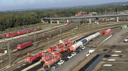Weitwinkelansicht eines großen Bahngeländes mit zahlreichen Gleisen, Oberleitungen, roten Lokomotiven und Güterwagen. Im Vordergrund stehen mehrere Fahrzeuge auf einer Straße neben den Gleisen. Im Hintergrund verläuft eine Brücke über die Bahnanlagen, dahinter sind Bäume und weitere Gleisfelder zu sehen.