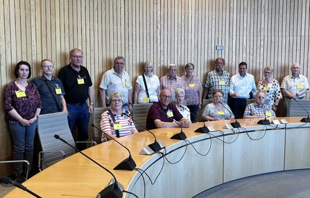 Gruppenfoto mit etwa 15 Personen in einem Besprechungsraum. Mehrere Personen stehen vor einer vertäfelten Holzwand, andere sitzen an einem großen gebogenen Konferenztisch mit Mikrofonen. Alle tragen gelbe Namensschilder.