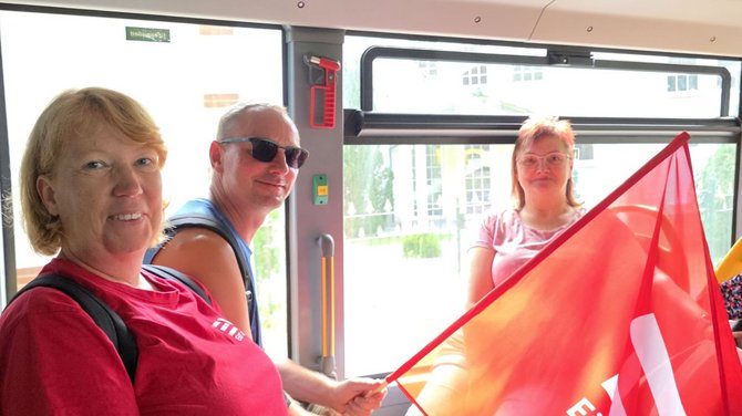 Drei Personen befinden sich im Innenraum eines Fahrzeugs mit großen Fenstern. Eine Person hält eine rote Fahne mit weißem EVG-Schriftzug. Durch die Fenster sind Gebäude und Vegetation erkennbar. Die Aufnahme ist durch das helle Tageslicht im Hintergrund stark aufgehellt.