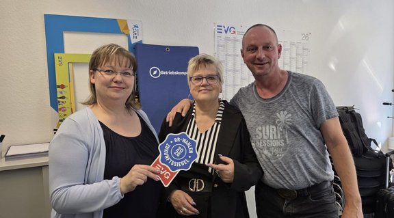 Drei Personen stehen in einem Büroraum nebeneinander. Eine Person links hält ein Schild mit der Aufschrift „BR‑Wahl Zukunftssiegel 2026 – EVG“. Im Hintergrund sind ein blauer Aufsteller mit dem Wort „Betriebsrat“ sowie Wandkalender und EVG‑Logos zu sehen. Rechts steht ein Rucksack auf einem Tisch oder Schrank.