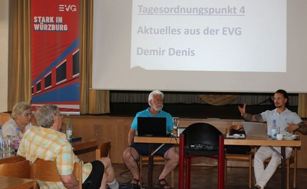 Mehrere Menschen sitzen in einem Raum an Tischen und hören einer Präsentation zu. Vorne sitzen zwei Personen mit Laptops an einem Tisch, dahinter ist eine Leinwand mit dem Text „Tagesordnungspunkt 4 Aktuelles aus der EVG Demir Denis“. Links steht ein rotes Banner mit der Aufschrift „STARK IN WÜRZBURG“.
