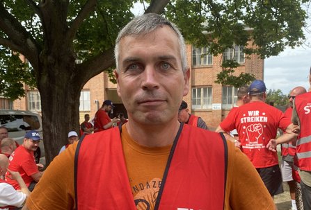 Porträt einer Person in einer roten Warnweste und orangefarbenem T-Shirt im Vordergrund. Im Hintergrund stehen und sitzen weitere Personen in roten Westen und roten T-Shirts. Sichtbare Aufschriften sind unter anderem „EVG“ und „WIR STREIKEN“. Dahinter befinden sich Bäume und Backsteingebäude.