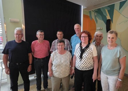 Gruppe von neun Personen steht nebeneinander in einem Innenraum vor einer Wand mit Muster. Die Personen schauen in die Kamera, einige tragen Brillen. Unterschiedliche Kleidung in gedeckten Farben, im Hintergrund ein dunkler Vorhang und Wandgestaltung.
