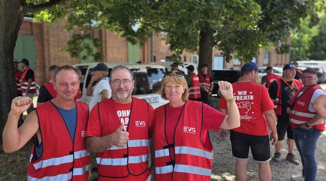 Drei Personen in roten EVG-Westen stehen im Vordergrund unter Bäumen. Zwei Personen heben einen Arm, eine Person zeigt einen erhobenen Daumen. Im Hintergrund befinden sich weitere Personen in roten Westen und T-Shirts, geparkte Fahrzeuge sowie Backsteingebäude.