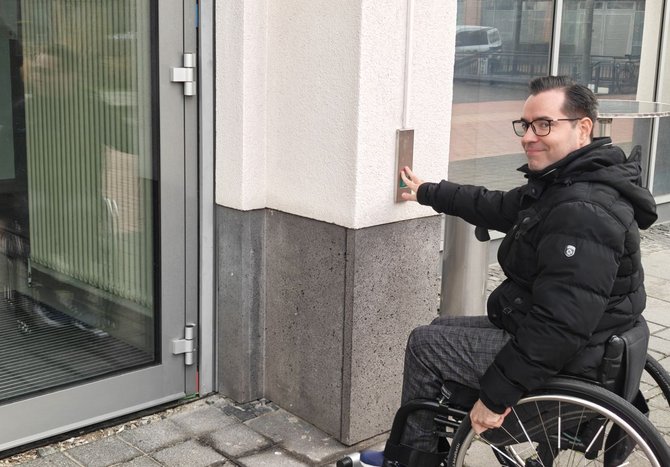 Eine Person sitzt in einem Rollstuhl neben dem Eingang eines Gebäudes und berührt einen Bedienknopf an einer Metallsäule neben einer Glastür. Der Eingangsbereich ist mit Steinplatten gepflastert, daneben befinden sich Fenster und Gebäudewände.