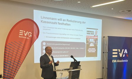 Vortragsraum mit einer Person an einem Rednerpult vor einer Projektionsfläche. Links steht ein rotes EVG-Banner mit der Aufschrift „EVG kommt an.“. Auf der Leinwand steht als Titel „Linnemann will an Reduzierung der Kassenzahl festhalten“. Rechts ist ein blaues Banner mit der Aufschrift „EVA Akademie“ zu sehen. Vor der Leinwand stehen ein Laptop, Mikrofone und ein Glas Wasser.