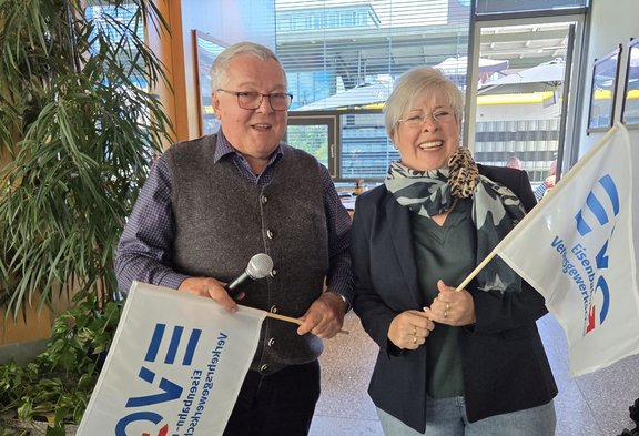 Zwei Personen stehen nebeneinander in einem Innenraum mit großen Glasfenstern und Pflanzen im Hintergrund. Beide halten weiße Fahnen mit blau-rotem Logo und dem gut lesbaren Text „Eisenbahn- und Verkehrsgewerkschaft.“ Die Person links hält zusätzlich ein Handmikrofon. Die Personen tragen Alltagskleidung mit Jacken und Oberteilen; eine Person trägt einen Schal. Im Hintergrund sind Sitzgelegenheiten, weitere Personen und ein moderner Gebäudekomplex durch die Fenster zu sehen.