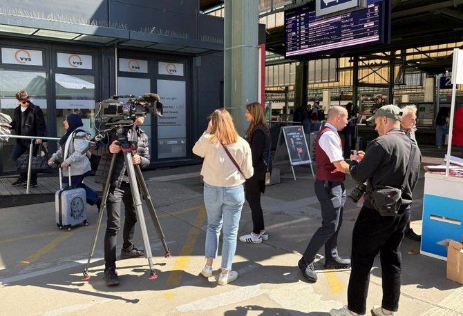 Eine größere Szene im Bahnhofsbereich mit mehreren Personen. Links steht eine Kamera auf einem Stativ, bedient von einer Person. Weitere Personen stehen in Gruppen zusammen. Im Hintergrund sind Schalter mit der Aufschrift „vvs“, Anzeigetafeln mit Zug- oder Abfahrtsinformationen sowie Bahnsteige und Gleise zu sehen.