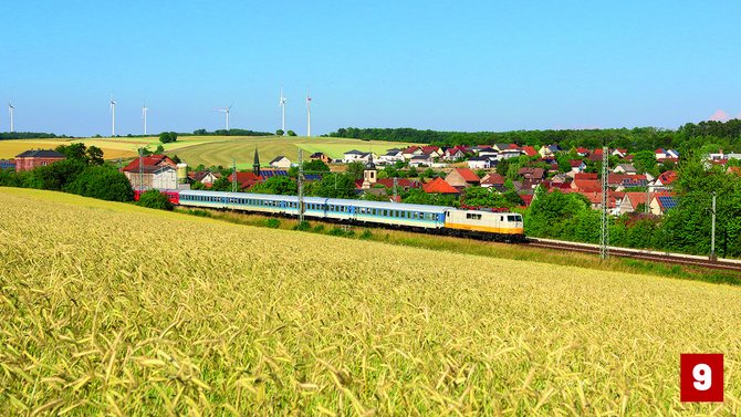 Heller Reisezug mit cremefarbener Lokomotive und mehreren blau-weißen Personenwagen auf einer Bahnstrecke zwischen einem goldgelben Feld und einem Dorf mit roten Dächern. Im Hintergrund stehen Windkraftanlagen auf sanften Hügeln. Unten rechts befindet sich eine rote Markierung mit der Zahl „9“.