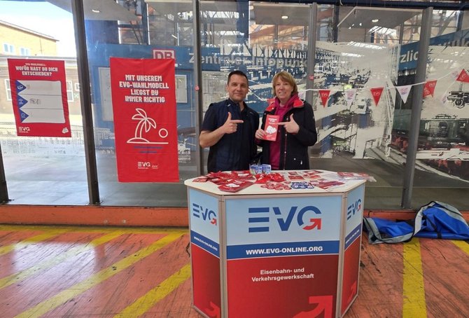 Zwei Personen stehen hinter einem kleinen, rechteckigen Informationsstand mit EVG‑Logo und der Aufschrift „EVG – Eisenbahn- und Verkehrsgewerkschaft“ sowie der Webadresse „www.evg-online.org“. Auf der Standfläche liegen rote und blaue Flyer, Aufkleber und weiteres Informationsmaterial. Beide Personen halten Daumen hoch. Hinter dem Stand befindet sich eine Glaswand mit Blick in eine große Industriehalle mit Maschinen und Fördertechnik. An der Glaswand hängen rote EVG‑Plakate mit den Texten „Wofür hast du dich entschieden?“ und „Mit unserem EVG‑Wahlmodell liegst du immer richtig“. Der Boden ist farbig markiert, rechts liegt eine Tasche am Boden.
