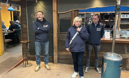 Innenbereich mit Holzwänden und einer Theke im Hintergrund. Drei Personen stehen nebeneinander vor der Theke; eine Person hält ein Mikrofon. Zwei Personen tragen dunkle Jacken mit der Aufschrift „EVG BERLIN“. Rechts im Vordergrund steht ein hoher, silberfarbener Heizpilz. Links sind Sitzplätze mit weiteren Personen an Tischen zu sehen. An der Wand befindet sich eine dunkle Tafel mit teilweise lesbarer Kreideschrift.