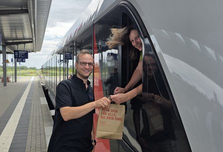 in Mann steht auf einem Bahnsteig neben einem modernen Zug und hält eine Papiertüte, während eine Person aus dem geöffneten Zugfenster herausgreift und etwas entgegennimmt.