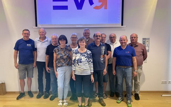 Dreizehn Personen stehen in zwei Reihen in einem hellen Raum vor einer Projektionsfläche. Zwei Personen stehen vorne mittig, neun weitere dahinter. Mehrere Personen tragen blaue Oberteile mit sichtbarem „EVG“-Logo. Im Hintergrund ist auf der Leinwand ein teilweise sichtbares großes „EVG“-Zeichen zu sehen. Der Boden ist aus hellem Holz, an den Seiten sind weiße Wände. Rechts an der Wand befindet sich ein Bedienfeld, links unten steht eine Papiertüte auf dem Boden.