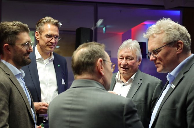 Fünf Personen stehen in einem Innenraum zusammen. Links stehen zwei Personen mit Brillen und Jackett. Dahinter steht eine weitere Person mit Jackett und Hemd. In der Bildmitte ist eine Person von hinten zu sehen, ebenfalls mit Jackett. Rechts steht eine weitere Person mit Brille und Jackett. Im Hintergrund sind Scheiben und Beleuchtung erkennbar.