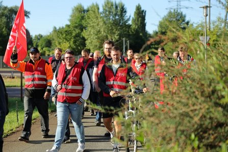 Eine Gruppe von Personen in roten Warnwesten geht auf einem asphaltierten Weg. Links trägt eine Person eine rote Fahne mit weißer Aufschrift. In der Mitte wird ein Fahrrad geschoben. Rechts befinden sich Büsche, im Hintergrund Bäume und Freiflächen bei Tageslicht.