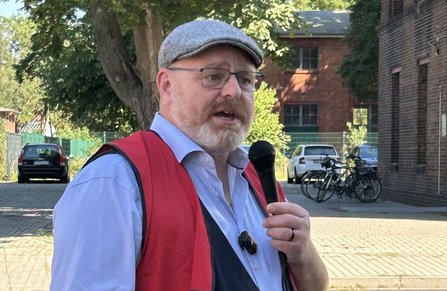 Person mit Schiebermütze, Brille, hellem Hemd und roter Weste hält ein Handmikrofon in der Hand und steht im Freien auf einem gepflasterten Hof. Im Hintergrund sind Bäume, Backsteingebäude, mehrere Fahrräder und geparkte Autos zu sehen.