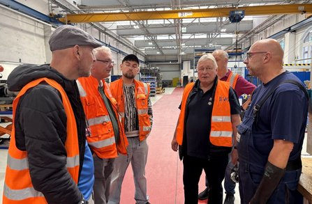 Sechs Personen stehen in einer großen Werkhalle mit rotem Boden zwischen Maschinen und Arbeitsbereichen. Vier Personen tragen orangefarbene Warnwesten mit Reflexstreifen, eine weitere Person trägt Arbeitskleidung mit Latzhose und Schutzhandschuh. Im Hintergrund sind ein gelber Hallenkran, Regale, Werkbänke und industrielle Anlagen zu sehen.