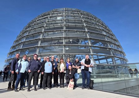Gruppe von Personen steht vor derselben gläsernen Kuppel aus einer etwas weiteren Perspektive. Mehrere Personen tragen Taschen oder Rucksäcke. Eine Stofftasche zeigt den Text „EVG BERLIN“. Die Kuppel füllt den oberen Bildbereich.