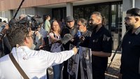 Mehrere Personen stehen im Freien vor einem Gebäude mit großen Fenstern und halten eine Pressebefragung ab. Im Vordergrund sind mehrere Mikrofone und ein Kamerateam mit einer Videokamera auf einem Schulterstativ zu sehen. Zwei Personen halten große ausgedruckte Fotos mit Porträtaufnahmen. Rundherum stehen weitere Personen, einige mit Kameras und Aufnahmegeräten, die auf die sprechenden Personen gerichtet sind. Das Licht ist hell und wirft deutliche Schatten.