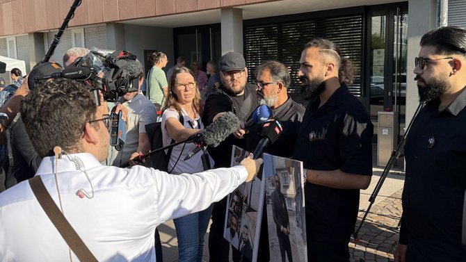 Mehrere Personen stehen im Freien vor einem Gebäude mit großen Fenstern und halten eine Pressebefragung ab. Im Vordergrund sind mehrere Mikrofone und ein Kamerateam mit einer Videokamera auf einem Schulterstativ zu sehen. Zwei Personen halten große ausgedruckte Fotos mit Porträtaufnahmen. Rundherum stehen weitere Personen, einige mit Kameras und Aufnahmegeräten, die auf die sprechenden Personen gerichtet sind. Das Licht ist hell und wirft deutliche Schatten.