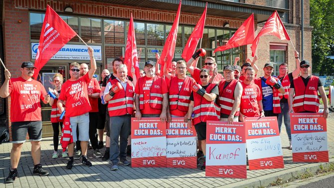 Größere Gruppe von Personen auf einem Gehweg vor einem Backsteingebäude mit Schaufenstern. Zahlreiche rote Fahnen mit EVG-Aufdruck werden hochgehalten. Mehrere rote Schilder tragen die Überschrift „MERKT EUCH DAS:“ und handschriftliche Texte, darunter „mehr Wertschätzung“, „Tarifverträge sind ein Muss!“ und „ohne EVG ohne UNS!“.