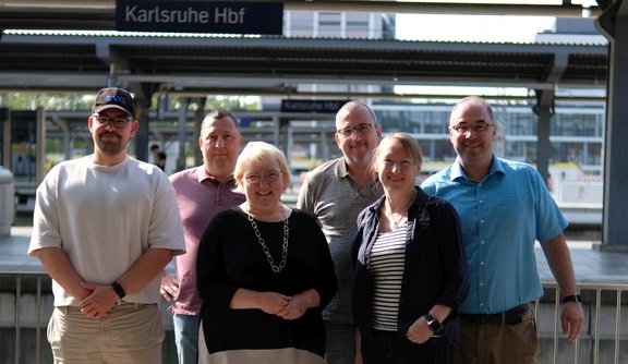 Sechs Personen stehen auf einem Bahnsteig vor dem Schild „Karlsruhe Hbf“ und blicken in die Kamera. Im Vordergrund stehen drei Personen, dahinter drei weitere. Eine Person trägt eine dunkle Kappe, eine andere ein hellblaues Kurzarmhemd. Im Hintergrund sind Bahnsteigdächer, Geländer, Gleisanlagen und weitere Schilder mit der Aufschrift „Karlsruhe Hbf“ zu sehen. Die Aufnahme entstand bei Tageslicht im Außenbereich eines Bahnhofs.