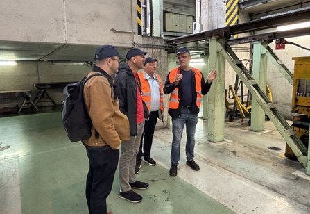 Vier Personen mit orangefarbenen Warnwesten und dunklen Kappen stehen in einer Industriehalle. Im Umfeld sind Stahlträger, eine Treppe, technische Anlagen und markierte Stützen sichtbar. Eine Person gestikuliert während die Gruppe daneben steht.