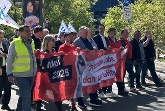 Eine größere Gruppe von Personen bewegt sich gemeinsam auf einer Straße. In der vorderen Reihe halten mehrere Personen ein breites rotes Banner. Auf dem Banner steht links „1. Mai 2026“ und rechts der Text „Erst unsere Jobs, dann eure Profite“, daneben ist ein DGB-Logo zu sehen. Einige Personen tragen Jacken, Westen oder Hemden, eine Person trägt eine gelbe Warnweste. Im Hintergrund sind weitere Personen mit weißen Fahnen mit blauen Streifen sowie Straßenschilder und Bäume zu erkennen.
