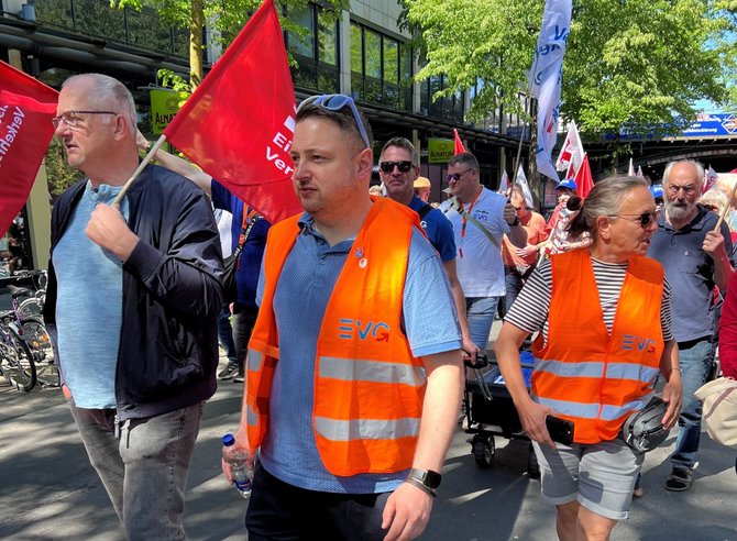 Nahaufnahme einer Gruppe gehender Menschen auf einer Straße. Im Vordergrund tragen mehrere Personen orangefarbene Warnwesten mit dem Aufdruck „EVG“. Eine Person hält eine rote Fahne mit weißem Schriftzug. Weitere rote und weiße Fahnen sind im Hintergrund zu sehen. Die Umgebung zeigt Bäume, Gebäude mit Glasfassaden und weitere Menschen, die sich in dieselbe Richtung bewegen.