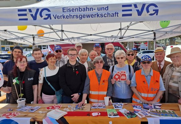 Unter einem weißen Pavillon mit der Aufschrift „EVG Eisenbahn- und Verkehrsgewerkschaft“ stehen mehrere Personen dicht nebeneinander hinter einem Holztisch. Auf dem Tisch liegen Flyer, Broschüren, Aufkleber, Anstecknadeln, ein Buch, ein Becher und kleine Gegenstände. Vor dem Tisch hängt eine Fahne in Schwarz‑Rot‑Gold. Einige Personen tragen orangefarbene Warnwesten mit „EVG“-Logo, andere Kleidung in verschiedenen Farben; einzelne tragen Kappen. Eine Person hält ein rotes, achteckiges Schild mit weißem Text, eine andere eine weiße Blume im Topf. Im Hintergrund sind Luftballons, ein rotes Fahrzeug und Gebäude im Außenbereich zu sehen.